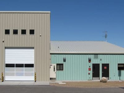 A small industrial building with beige and light green metal siding, a tall garage door, and multiple small windows under a clear blue sky.