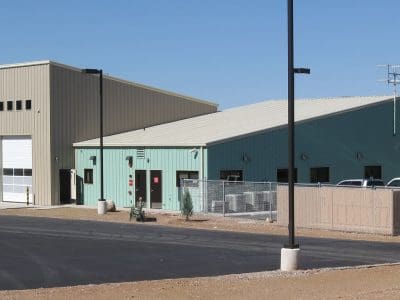 A light industrial building with tan and green metal siding, large garage door, small windows, fenced area, and a paved lot under clear skies.