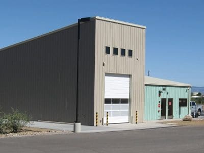 A beige industrial building with a tall garage door stands next to a smaller light green structure on a paved lot under a clear blue sky.