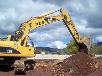 A yellow excavator is digging and moving soil at a construction site with mountains and cloudy sky in the background.