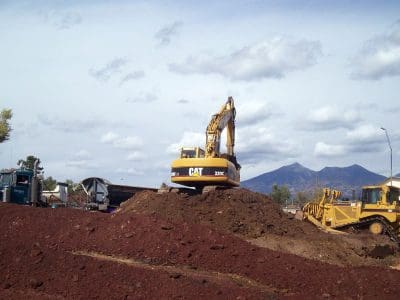 Construction site with a CAT excavator and bulldozer working on a large dirt mound, with a truck parked nearby and mountains in the background under a partly cloudy sky.
