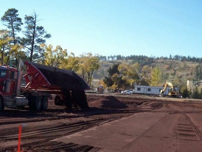 A red dump truck unloads soil at a construction site with machinery and houses in the background, surrounded by trees and hills.