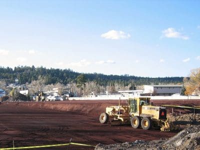 A yellow grader is parked on a large, freshly leveled dirt construction site, with buildings and trees in the background under a clear blue sky.
