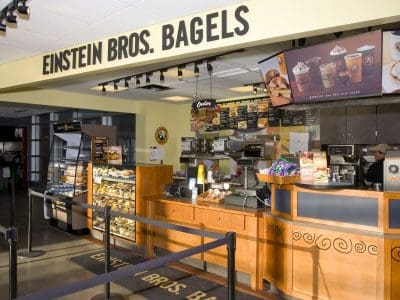 Einstein Bros. Bagels counter with pastries on display, digital menu boards, and ordering area in a well-lit cafe setting.