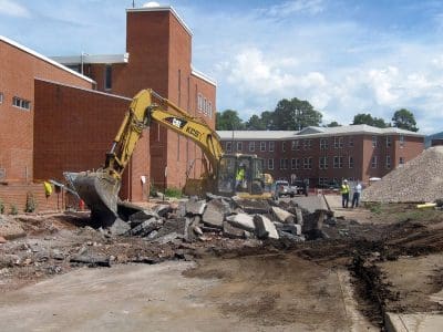 An excavator breaks up concrete outside a brick building while two workers stand nearby; piles of debris and dirt are visible on the construction site.