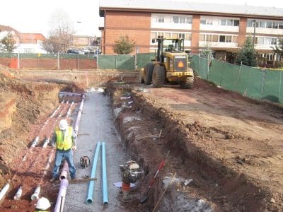 Construction site with a worker and pipes in a trench, a backhoe loader in the background, and a building behind a green fence.