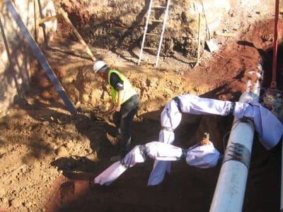 A construction worker wearing a safety vest and helmet stands in a dirt trench near insulated pipes and a ladder at an outdoor worksite.