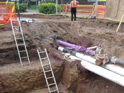 Open construction site with exposed underground pipes, three ladders in the trench, and a worker in a safety vest standing near fencing in the background.