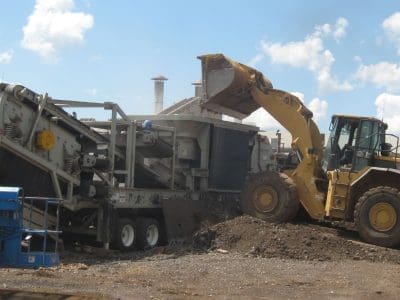 A large yellow front loader deposits soil into an industrial screening machine at a construction site under a partly cloudy sky.