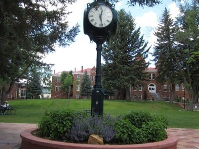 A black clock stands in a circular brick planter surrounded by greenery, with red brick campus buildings and trees in the background.