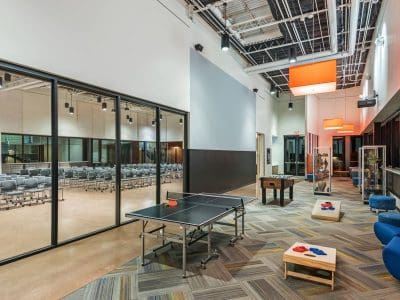 Modern recreation area with a ping pong table, foosball table, seating, and large windows overlooking an auditorium with rows of chairs.