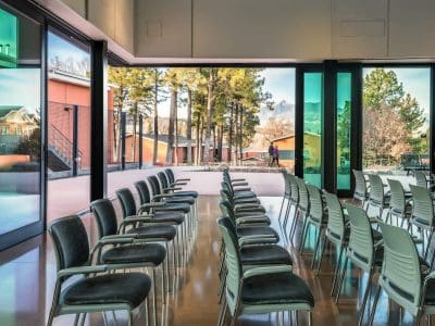 Rows of empty chairs are arranged in a room with large glass doors open to an outdoor area with trees, buildings, and a person walking in the distance.