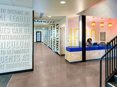 A modern office lobby with a reception desk, wall art with motivational text, a staircase, and bright lighting. A person is seated at the reception desk.