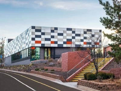 A modern building with a checkered facade in black, white, and orange, next to a road with a yellow-striped stairway and landscaped surroundings.