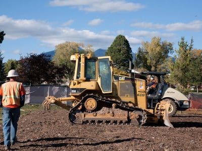 Two construction workers wearing safety gear stand near a yellow bulldozer and a roller on a dirt lot with trees and fencing in the background.