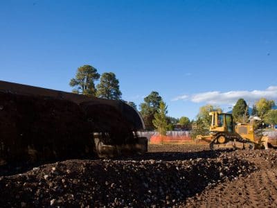 A construction site with a yellow bulldozer and a dump truck moving dirt, surrounded by trees and temporary orange fencing under a clear blue sky.