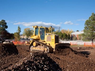 A yellow bulldozer moves dirt on a construction site, with trees, buildings, and orange safety fencing in the background under a blue sky.