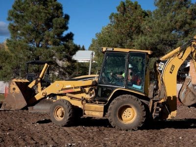 A yellow backhoe loader is parked on a dirt construction site, surrounded by trees and buildings in the background under a clear sky.