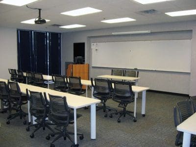 Empty classroom with rows of tables and chairs, a large whiteboard at the front, and a podium with computer equipment near the corner.