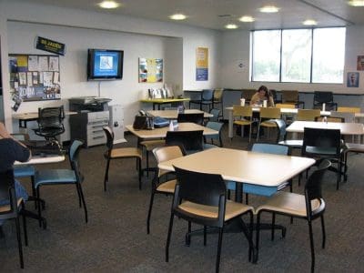 A break room with several tables and chairs, a large window, wall posters, a TV, and a few people sitting and working or resting.