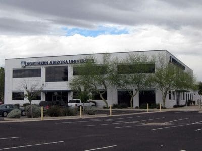A white, two-story building with "Northern Arizona University" signage, surrounded by trees and an adjacent parking lot with several parked cars.