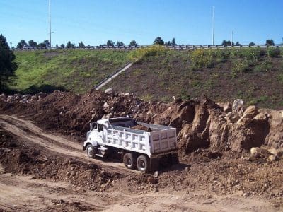 A white dump truck is parked in a dirt construction site near an embankment, with a grassy slope and a road in the background.