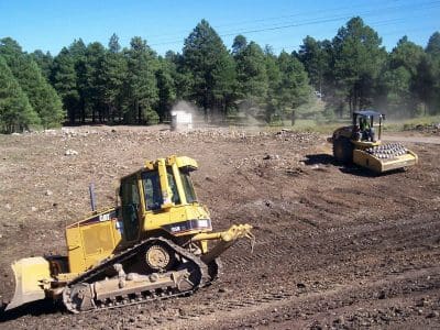 A yellow bulldozer and a soil compactor work on clearing and leveling dirt at a construction site near a forested area.