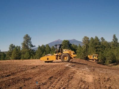 A yellow soil compactor and bulldozer work on a cleared dirt area surrounded by pine trees with mountains in the background under a clear blue sky.