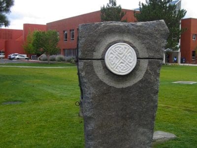 A large stone sculpture with a circular white emblem stands on a grassy lawn, with a red brick building and trees in the background.