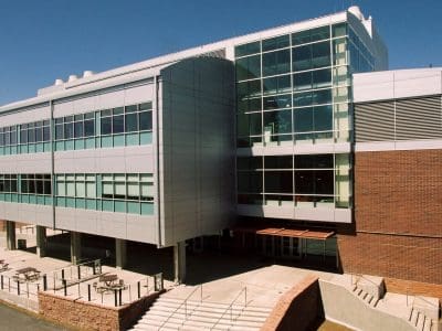 Modern three-story building with large windows and brick accents, featuring a main entrance with stairs and a ramp under a clear blue sky.