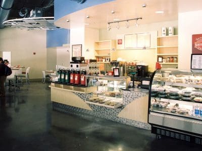 Self-service coffee and food counter with refrigerated display case and several beverages, located in a modern, well-lit cafe with a few people in the background.