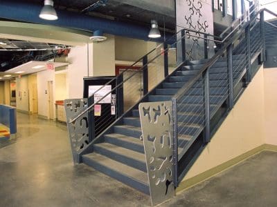 A modern indoor staircase with metal railings and decorative panels, located in a brightly lit building with polished concrete floors and colored tile accents.