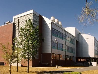Modern academic building with a mix of brick and metal exterior, large windows, and rooftop ventilation units, surrounded by trees and a paved walkway.