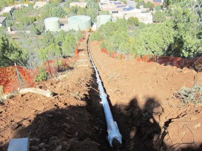 A pipeline installation runs down a dirt slope with orange safety fencing on both sides; storage tanks and buildings are visible in the background.