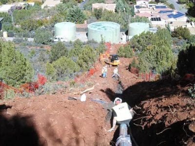 Workers install a large pipeline in a deep trench on a hillside, with residential buildings and water tanks visible in the background.