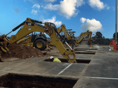 Several excavators are parked near rectangular pits in the ground, with a construction worker in a hard hat working inside one pit, and equipment lined up along a paved surface.