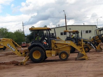 Several yellow backhoe loaders are parked on a dirt lot near a beige industrial building under a cloudy sky.
