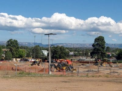 A construction site with several vehicles and equipment on dirt ground, surrounded by fencing, with trees and mountains in the background.