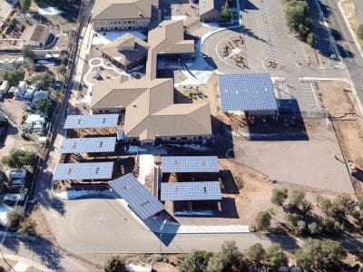 Aerial view of a large building complex featuring the Payson USD PV System with several solar panel structures in the parking area, surrounded by roads, trees, and adjacent smaller buildings.
