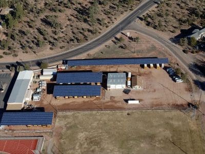 Aerial view of a facility with multiple buildings and parking areas, featuring the Payson USD PV System with solar panels on rooftops, surrounded by roads, trees, and an adjacent athletic field.