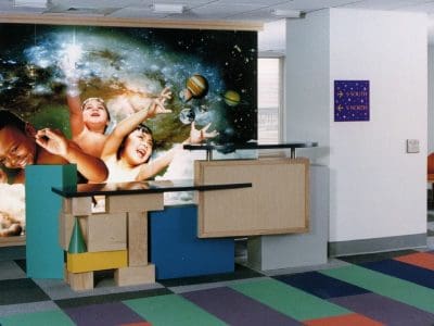 A children's play area with colorful tiles, soft chairs, geometric blocks, and a mural of kids reaching for planets and stars.