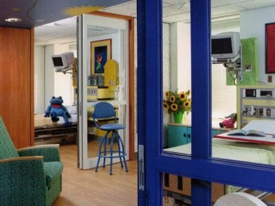 A brightly decorated hospital room with medical equipment, a hospital bed, a green armchair, sunflowers, colorful artwork, and a blue stuffed toy on another bed.