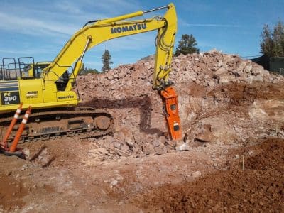 A yellow Komatsu excavator uses a hydraulic breaker attachment to break rocks at a construction site, with piles of rubble and soil in the background.