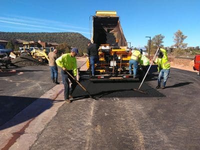 Construction workers in safety gear spread asphalt on a road while a dump truck unloads material and machinery operates in the background under a clear sky.