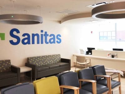 A modern medical clinic waiting area with gray and yellow chairs, a Sanitas logo on the wall, and a reception desk in the background.
