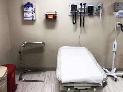 An empty medical exam room with an exam table, medical instruments on the wall, a trash can, and various supplies.