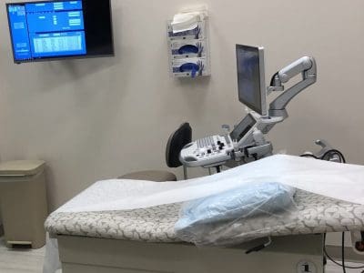 A medical examination room with an exam table, an ultrasound machine, a wall-mounted monitor displaying data, and medical supplies on the wall.