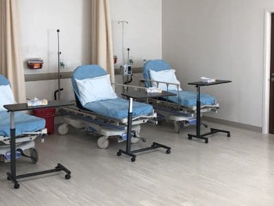 Three empty hospital beds with blue sheets and white pillows are lined up in a clean, well-lit room with medical equipment and privacy curtains.
