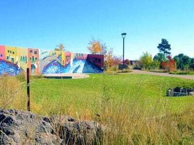 A colorful mural featuring buildings and water is painted on a wall beside a grassy area with a walkway, trees, and a bench under a clear blue sky.