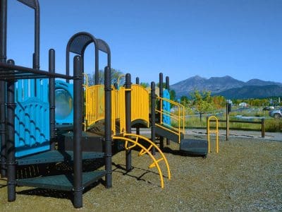 A modern playground with blue and yellow equipment sits on a gravel surface, with mountains and trees visible in the background under a clear blue sky.